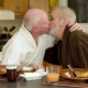 Two older men kissing in kitchen at breakfast